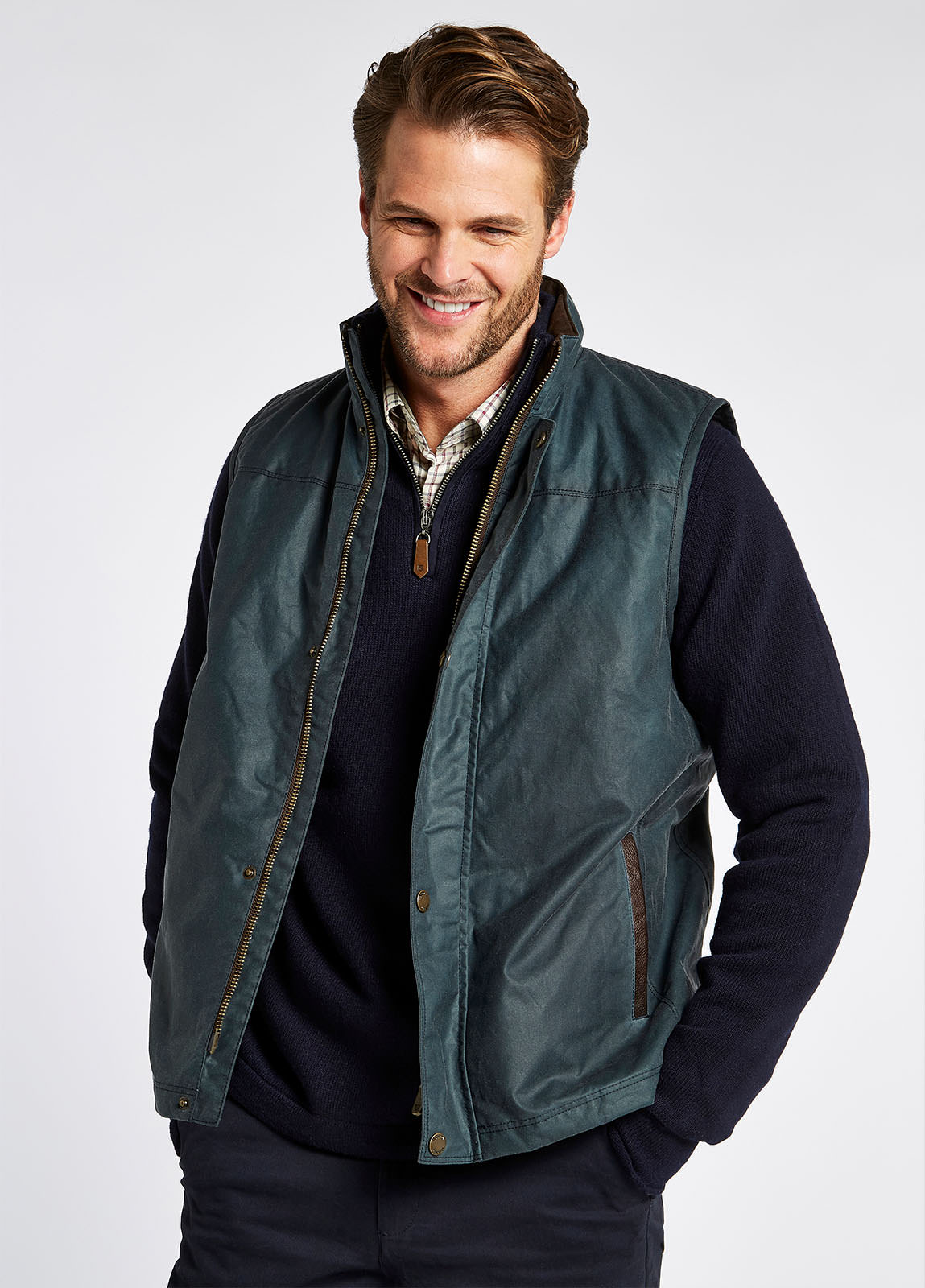 Waist up view of a male model wearing a Dubarry Mayfly wax gilet jacket for men. dark pebble blue green coloured gilet with full zip opened with buttons along the zip. The model is also wearing a dark jumper and light shirt underneath the gilet.